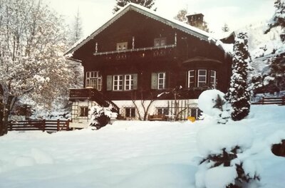 Zweistöckiges Holzhaus mit Balkon und weißem Sockel in einer verschneiten Gartenlandschaft.