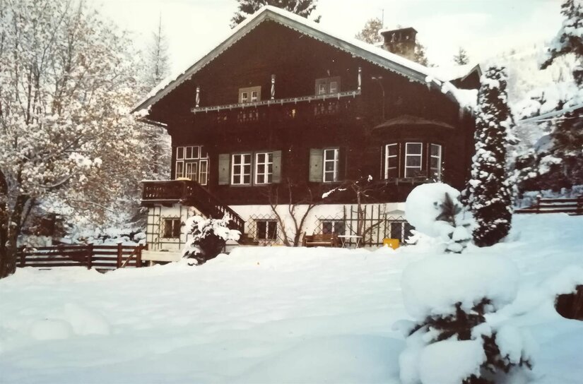 Villa Momo im Winter Zweistöckiges Holzhaus mit Balkon und weißem Sockel in einer verschneiten Gartenlandschaft.