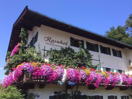 Außenansicht der Pension Rainhof mit blumengeschmücktem Holzbalkon und grünen Fensterläden vor blauem Himmel.