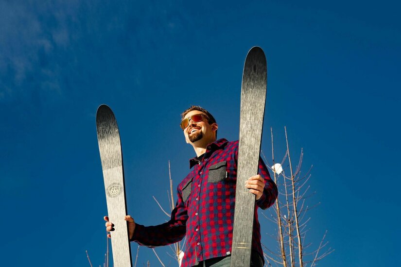 Ein Mann im karierten Hemd hält zwei dunkle Skier senkrecht vor blauem Himmel.