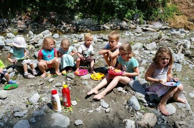 Eine Gruppe Kinder sitzt am Ufer eines kleinen Baches und genießt ihr Essen. Sie haben bunte Teller und scheinen eine schöne Zeit in der Natur zu verbringen.