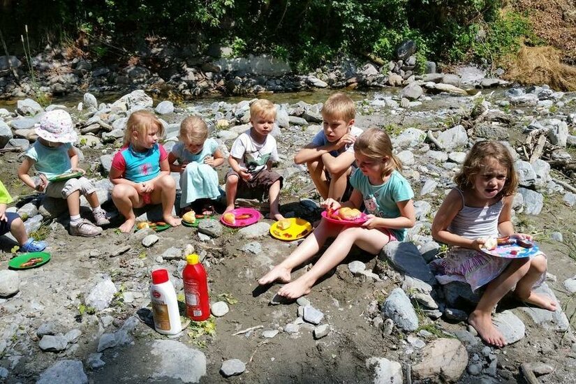 Eine Gruppe Kinder sitzt am Ufer eines kleinen Baches und genießt ihr Essen. Sie haben bunte Teller und scheinen eine schöne Zeit in der Natur zu verbringen.