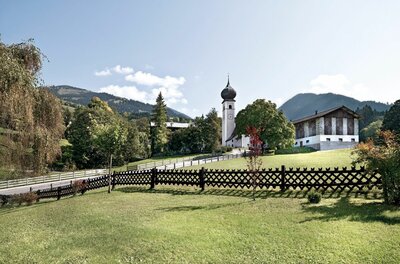 Ortsansicht Aurach bei Kitzbühel Kirche mit Zwiebelturm und Bauernhaus hinter einem Holzzaun auf einer grünen Wiese vor Bergkulisse.