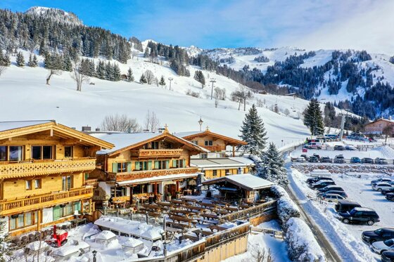 Verschneite Holzhütten mit Außenterrasse vor bewaldeten Berghängen im Winter.