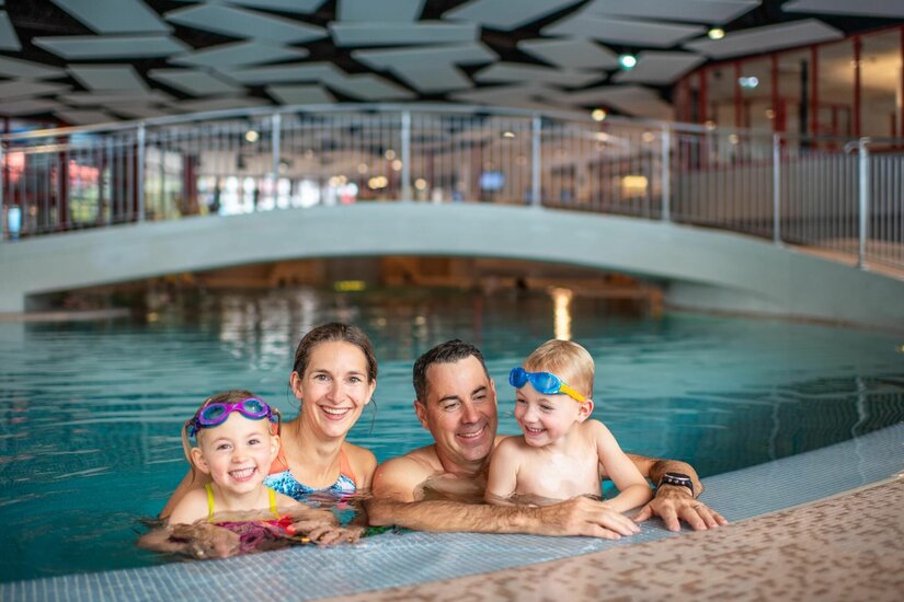 Eine vierköpfige Familie mit zwei Kindern im Wasser eines Hallenbades vor einer Brücke.