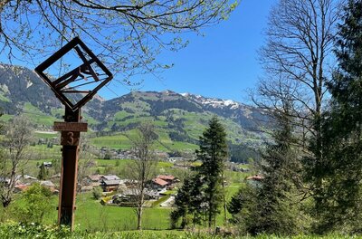 Ein hölzernes Kreuzweg-Relief im Vordergrund vor einer alpinen Tal- und Berglandschaft bei blauem Himmel.