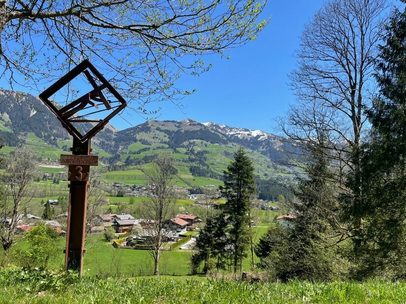 Station am Kreuzweg Ein hölzernes Kreuzweg-Relief im Vordergrund vor einer alpinen Tal- und Berglandschaft bei blauem Himmel.