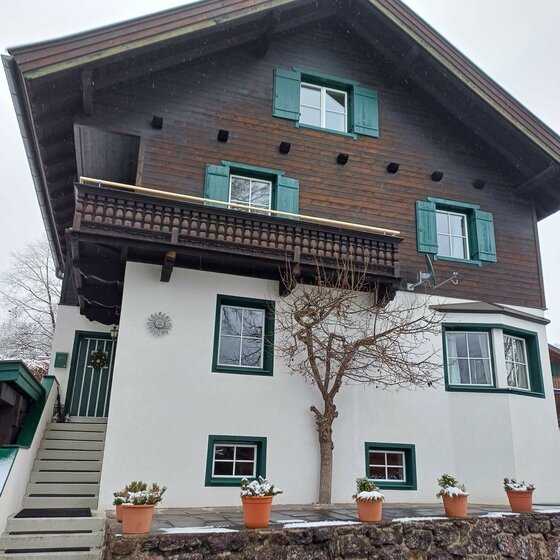 Alpines Wohnhaus mit weißer Fassade, dunklem Holzobergeschoss, grünem Fensterladen und Balkon.