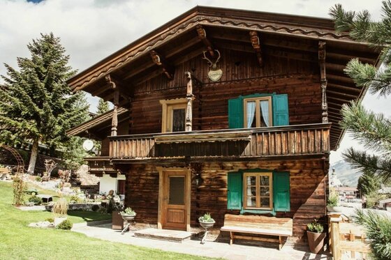 Jagahäusl Jochberg Außenansicht Zweistöckiges Holzhaus mit Balkon, grünen Fensterläden und einer Holzbank davor im Garten.