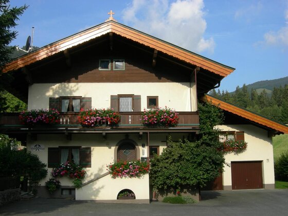 Zweistöckiges Alpenhaus mit Holzbalkon, Blumenschmuck und Satteldach vor bewaldetem Berg.