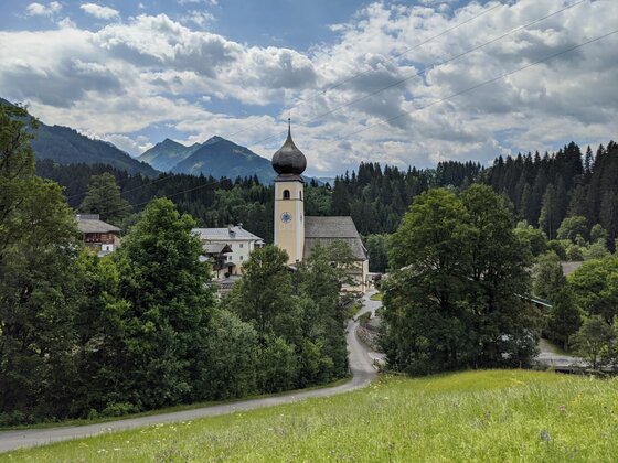 Kirche in Aurach bei Kitzbühel Kirche mit Zwiebelturm in einer alpinen Landschaft mit bewaldeten Bergen unter bewölktem Himmel.