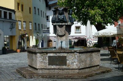 <p>Ein schöner Brunnen auf einem gemütlichen Platz. Am Brunnen sind Margarethe Maultasch, Ludwig der Strenge und Kaiser Maximilian abgebildet, umgeben von Gebäuden und Terrassen mit Sonnenschirmen.</p>