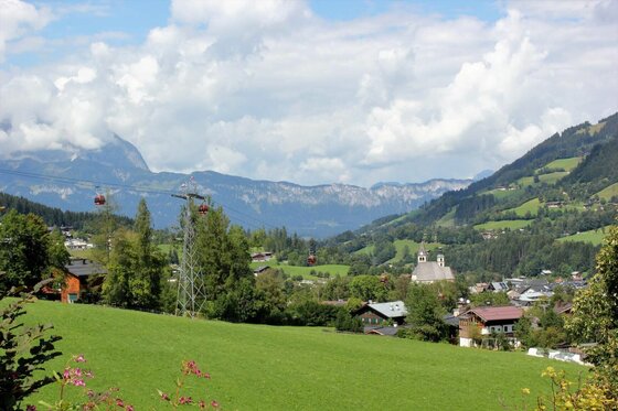 Aussicht auf Kitzbühel Blick über eine grüne Wiese auf ein Alpendorf mit Seilbahn und Bergen im Hintergrund.