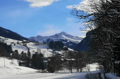 Verschneites Bergdorf mit Kirchturm vor schneebedeckten Berggipfeln unter blauem Himmel.