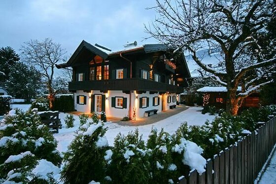 Winter am Weberhaus Beleuchtetes zweistöckiges Alpenhaus mit dunklem Holzobergeschoss in einer verschneiten Gartenanlage bei Dämmerung.
