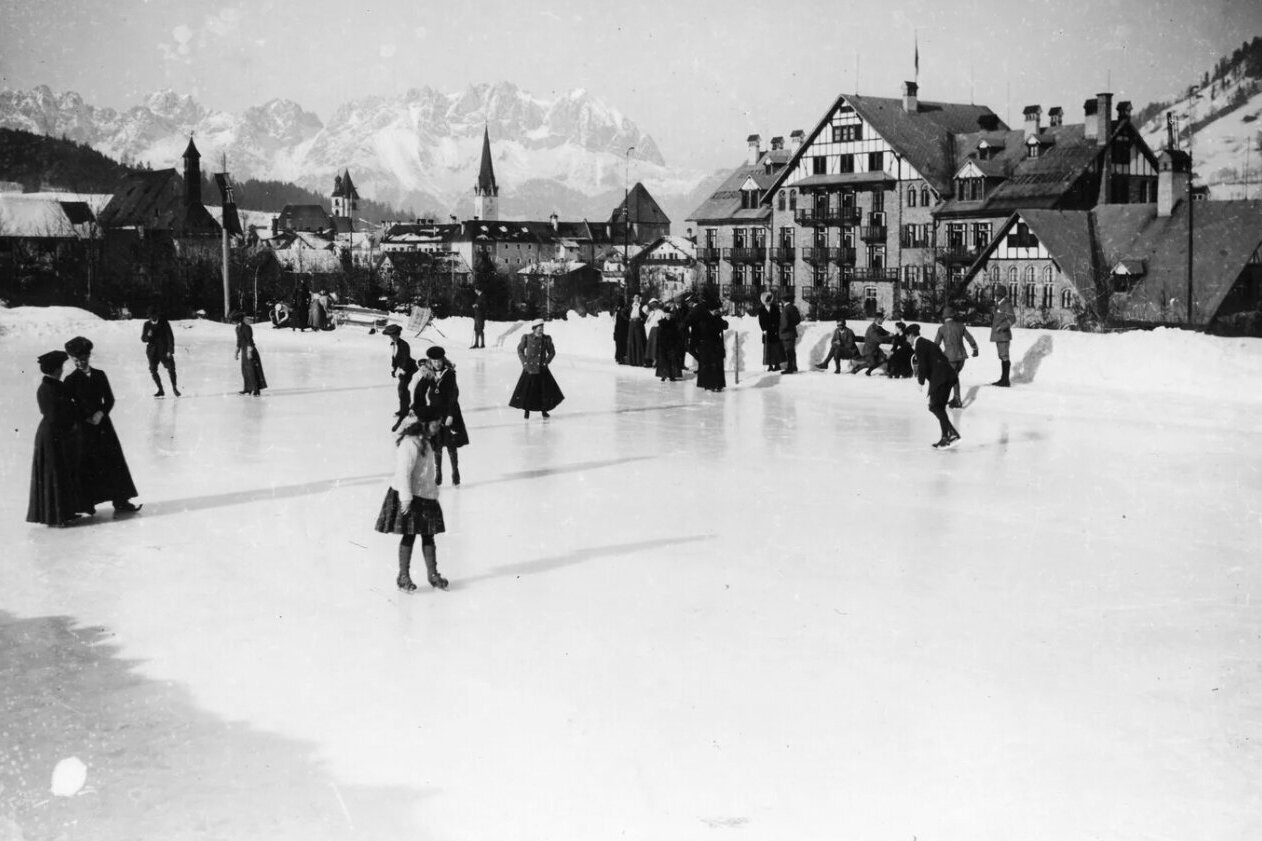 Women and men ice skating behind the Grand Hotel Kitzbühel, with the Wilde Kaiser visible in the background