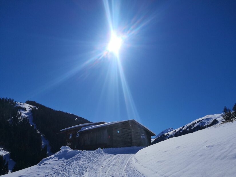 Holzhütte in verschneiter Berglandschaft unter strahlend blauem Himmel mit Gegenlicht.
