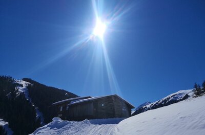 Holzhütte in verschneiter Berglandschaft unter strahlend blauem Himmel mit Gegenlicht.