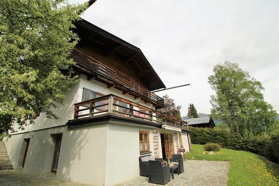 Lodge 242 Apartment Lebenberg Zweistöckiges alpines Gebäude mit Holzfassade, Balkon und einer Steinterrasse mit Sitzmöbeln.