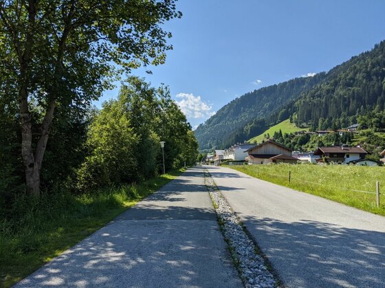 Radweg Kitz-Connection Asphaltierter Rad- und Wanderweg neben einer Straße in einem Tal mit bewaldeten Bergen im Hintergrund.
