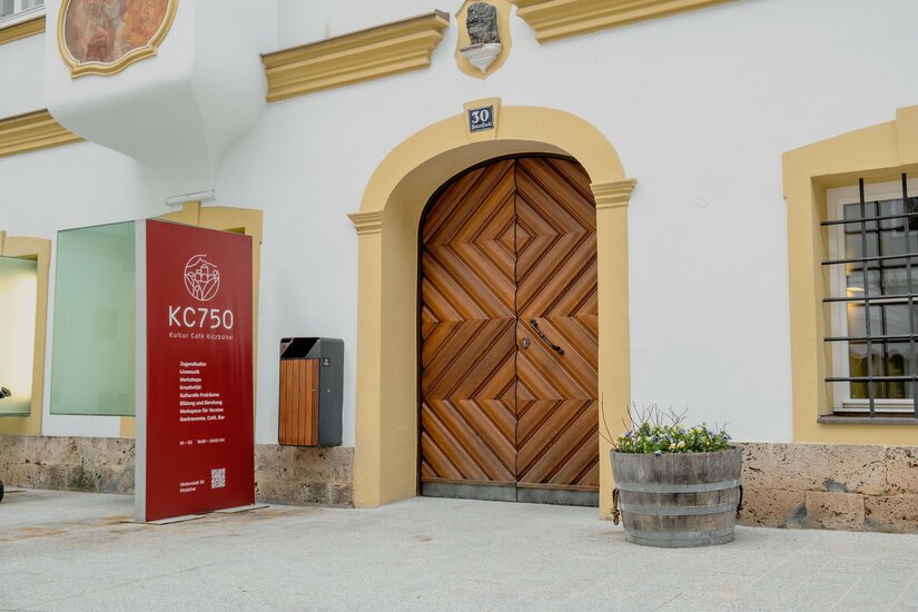 Eingangsbereich des KC750 Kultur Café mit großem Holztor und roter Informationstafel.