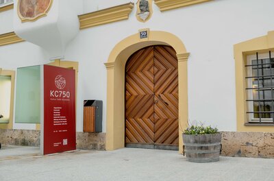 Eingangsbereich des KC750 Kultur Café mit großem Holztor und roter Informationstafel.