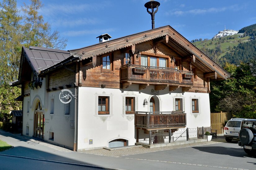 Alpines Wohnhaus mit weißer Fassade, Holzbalkonen und Satteldach vor bewaldeten Bergen.