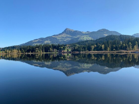 Sommer am Bergsee Ein ruhiger Bergsee mit Spiegelung der umliegenden Nadelwälder und Berggipfel unter blauem Himmel.