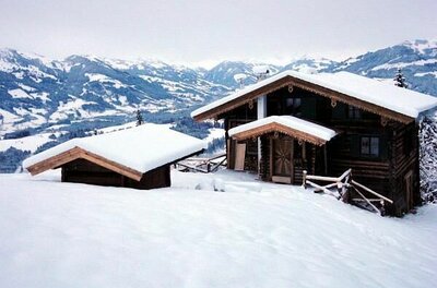 Verschneite Holzhütte und ein Nebengebäude in einer alpinen Winterlandschaft mit Bergen im Hintergrund.