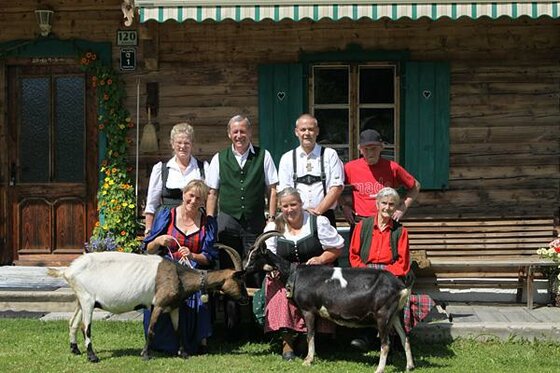 Familie vor dem Kainzergütl Eine Gruppe von sechs Personen in Tracht steht mit zwei Ziegen vor einem Holzhaus.