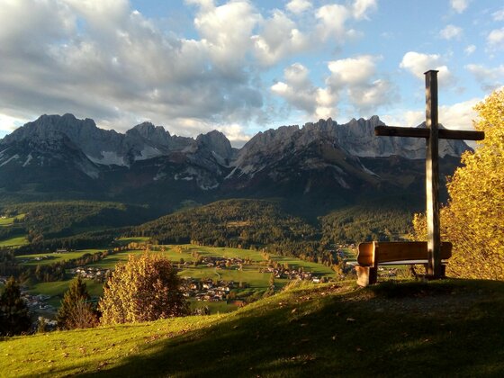 Eine malerische Berglandschaft mit hohen Gipfeln und buntem Herbstwald. Im Vordergrund steht ein Holzbank mit einem Kreuz, das zur Aussicht einlädt.