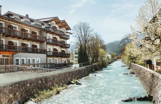 HENRI Country House Kitzbühel Mehrstöckiges Hotelgebäude mit Holzbalkonen direkt an einem befestigten Flusslauf in einer Bergregion.