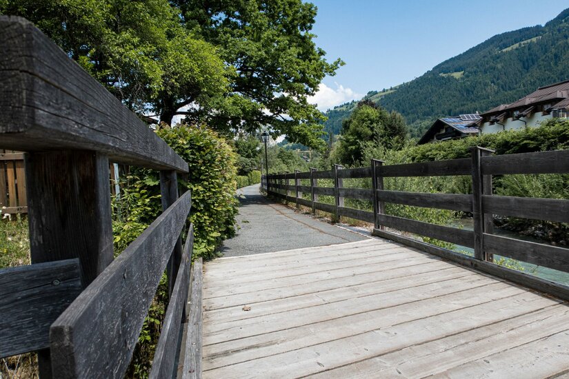 Holzbrücke führt auf einen asphaltierten Wanderweg entlang eines Zauns vor bewaldeten Bergen.