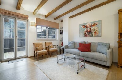 Wohnbereich Bacherwiese Penthouse Wohnzimmer mit grauem Sofa, zwei Sesseln, Holzbalkendecke und einem Kuhgemälde an der Wand.