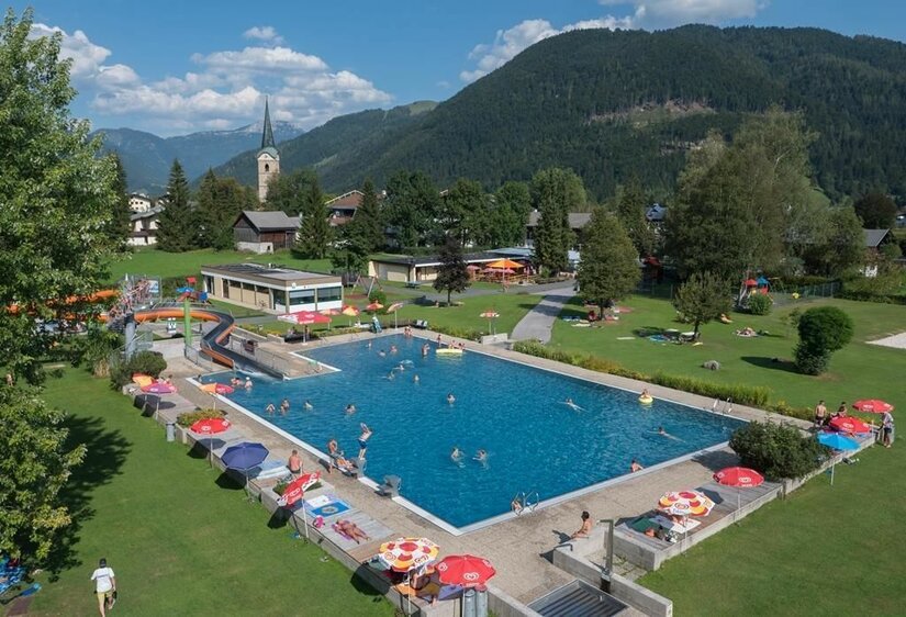 Freibad I Ein großer Swimmingpool umgeben von grünen Wiesen und Bäumen. Kinder und Erwachsene genießen das Wasser an einem sonnigen Tag.