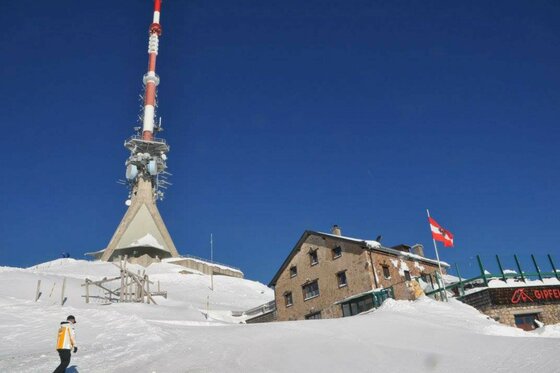 Ein rot-weißer Sendemast und ein steinernes Gipfelhaus in einer verschneiten Gebirgslandschaft unter blauem Himmel.