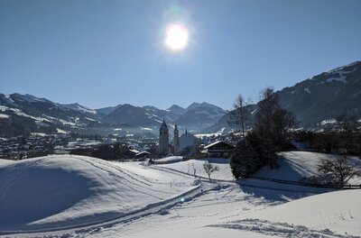 Verschneite Hügellandschaft mit Blick auf ein Dorf mit Kirchturm und umliegende Berge unter blauem Himmel.