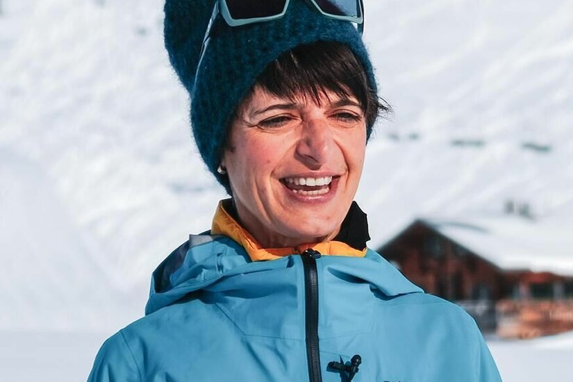Doris Ritter - Bikeguide Porträt einer Frau in blauer Outdoorjacke und Strickmütze vor verschneitem Berghang.
