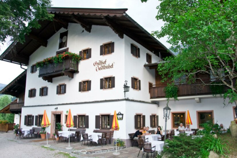 Traditioneller Gasthof mit weißer Fassade, dunklen Fensterläden und einer Außenterrasse mit Sonnenschirmen.