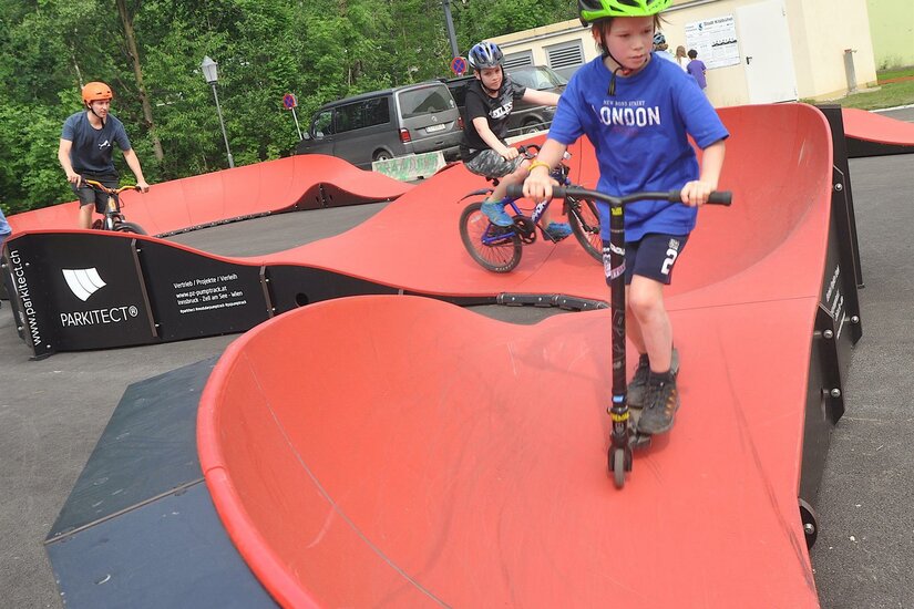 Ein Junge auf einem Roller und zwei Radfahrer befahren einen roten Pumptrack.