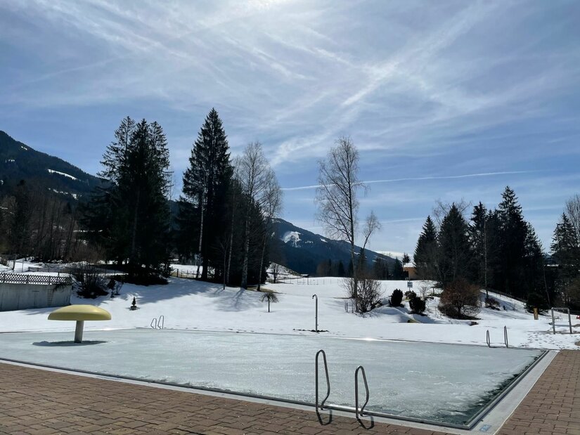 Freibad im Winter Ein zugefrorenes Freibadbecken in einer verschneiten Berglandschaft mit Nadelbäumen unter blauem Himmel.