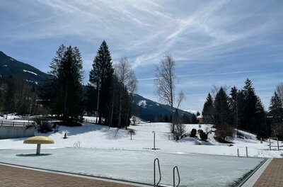 Freibad im Winter Ein zugefrorenes Freibadbecken in einer verschneiten Berglandschaft mit Nadelbäumen unter blauem Himmel.