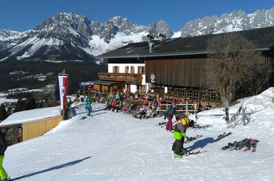 Eine alpine Skisportlandschaft mit schneebedeckten Bergen und einem Holzhaus im Hintergrund. Mehrere Skifahrer und Snowboarder genießen die Piste.