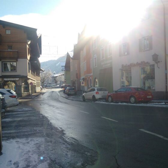 Blick entlang einer asphaltierten Straße in einem Alpendorf mit mehrstöckigen Häusern und parkenden Autos bei Gegenlicht.