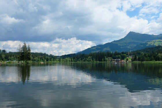 Hörpfing Runde am See Blick über einen ruhigen See auf bewaldete Ufer und einen markanten Berggipfel unter bewölktem Himmel.