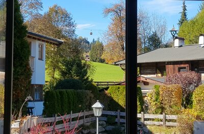 Blick auf die Gondel Blick durch ein Fenster auf alpine Häuser, Gärten und eine Seilbahngondel vor bewaldeten Hügeln.