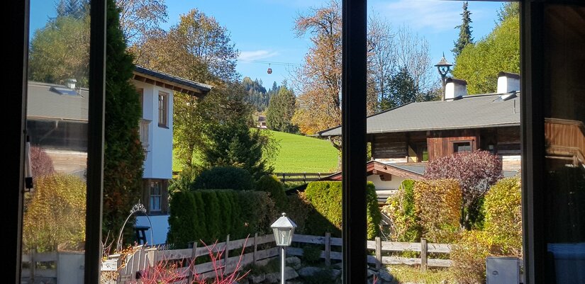 Blick durch ein Fenster auf alpine Häuser, Gärten und eine Seilbahngondel vor bewaldeten Hügeln.