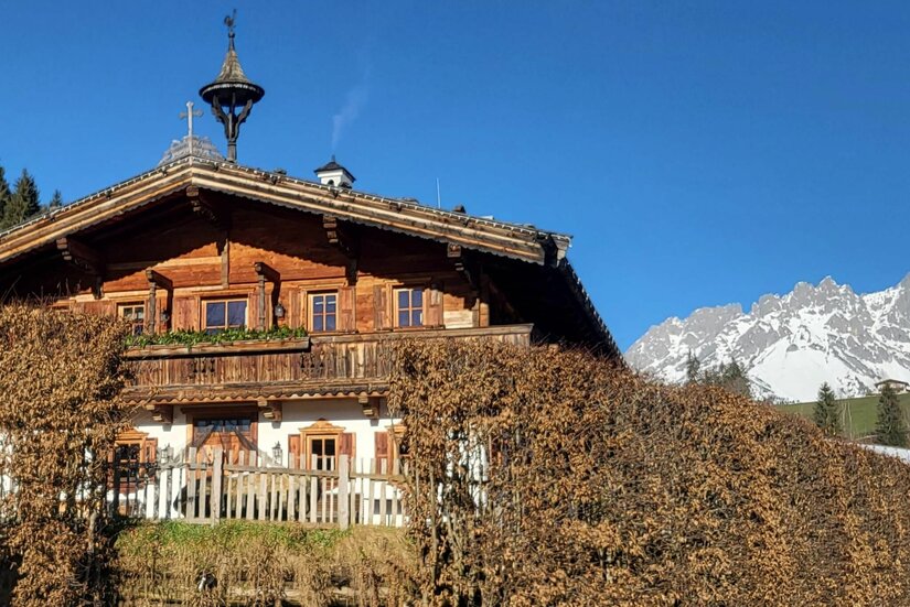 Traditionelles Holzhaus mit Balkon vor schneebedecktem Gebirgsmassiv unter blauem Himmel.