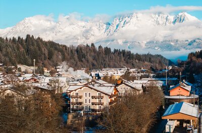 Blick über verschneite Hausdächer in Kitzbühel auf das schneebedeckte Massiv des Wilden Kaisers unter blauem Himmel.