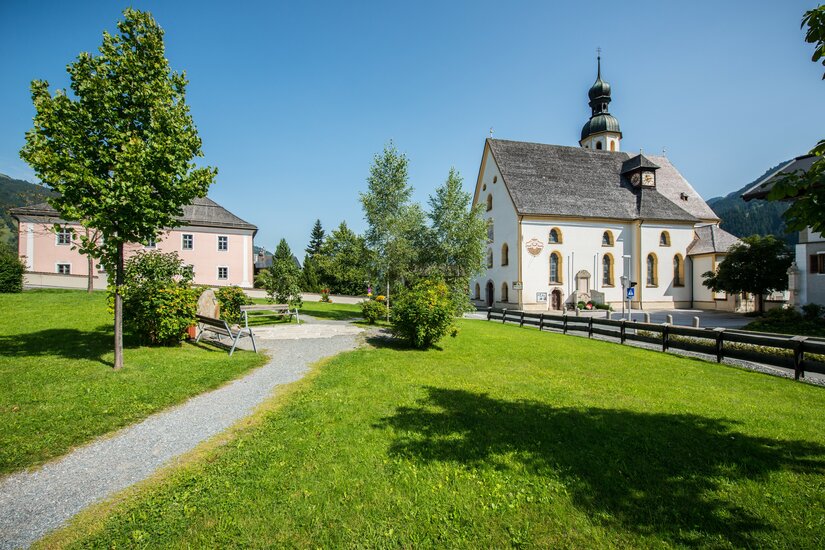 Weiße Dorfkirche mit Zwiebelturm neben einer Grünanlage mit Kiesweg und Parkbänken in Jochberg.